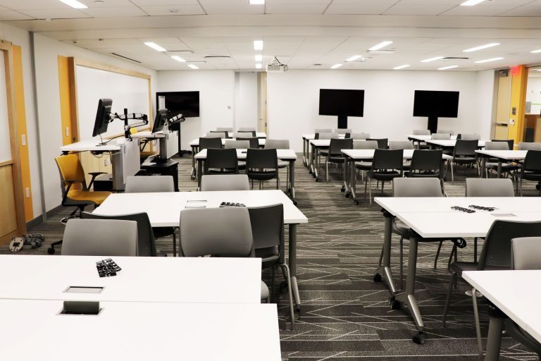 Classroom with tables surrounded by desks, with white boards and large monitors around the walls