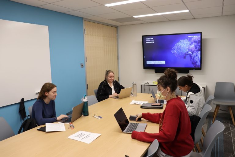 Four students sit around a conference table, each looking at laptop computers or paper notes.