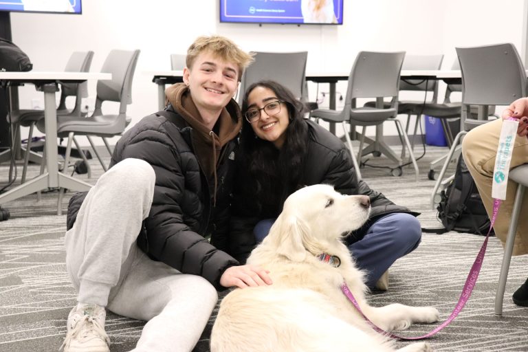 A young man and young woman sitting on the floor, petting a white golden retriever.