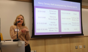 Woman standing at a podium in front of a projector screen that shows an agenda with the title Nancy Tannery Staff Development Day.