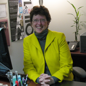 A white woman in a yellow blazer sitting behind her desk and smiling.