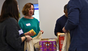People standing around a table that has boxes of individually packaged snacks. The people are holding canvas bags and placing snacks inside.