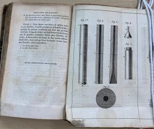 A book lying open on a table. The left page features text, while the right page features an illustration of the parts of Laennec's stethoscope, including the wood tube, the flared end, and the flat round base.