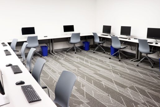 White tables wrap in a U-shape around a room, with Mac desktop computers set up at intervals, with a chair in front of each computer.