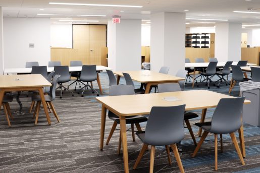 A large open room with several wood tables each with four chairs, and some longer white tables with six chairs. Bookshelves are visible in the background.