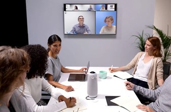 People sitting around a conference table. In the middle of the table is a cylindrical piece of technology, the Meeting Owl 3. On a screen behind them are faces of remote participants attending the meeting.