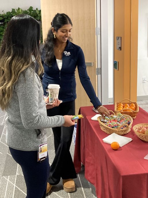 Two women, one wearing a sweater with a Pitt logo and the other with a UPMC badge, stand by a table with fruit and granola bars.