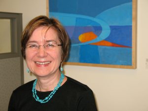 Gosia Fort, a white woman with short brown hair and glasses, pictured in front of a blue and orange painting.