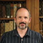 White man with brown and gray hair and beard, wearing a striped button down shirt, in front of a shelf of books.