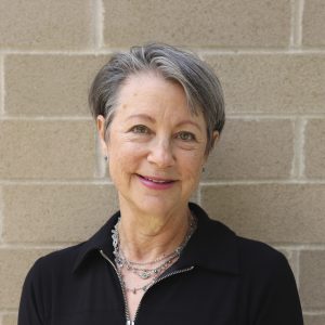A white woman with short gray hair, wearing a dark sweater, standing in front of a brick wall.