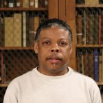 A BLack man with a mustache, wearing a white shirt, standing in front of a bookshelf.