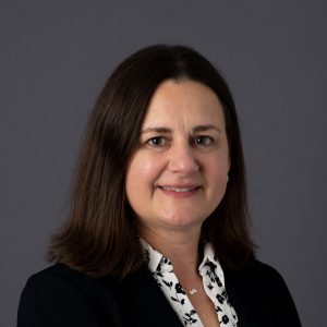 Headshot of a white woman with long brownhair, wearing a dark blazer over a white shirt.